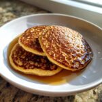 Three golden brown oatmeal pancakes stacked in a white bowl, drizzled generously with syrup.