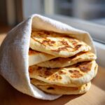 A stack of freshly cooked, fluffy pita bread wrapped partially in a white cloth, showing golden brown spots.