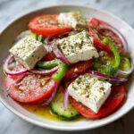 Vibrant close-up of a fresh greek salad featuring thick slices of tomato, cucumber, red onion, green pepper, and large cubes of feta cheese drizzled with olive oil and herbs.