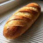 A freshly baked, golden-brown french bread loaf with crisp scoring resting on a wire cooling rack.