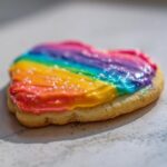 Close-up of a heart-shaped sugar cookie topped with vibrant, striped rainbow cookie icing and sparkling sugar crystals.