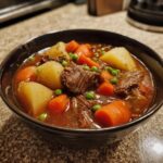Close-up of a dark bowl filled with rich vegetable beef soup, featuring tender beef chunks, carrots, potatoes, and peas.