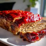Close-up of a juicy meatloaf topped with a thick, bright red glaze, with one slice cut and resting beside the main loaf.