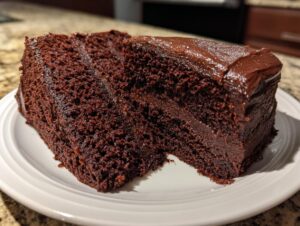 A thick, moist slice of homemade chocolate cake with rich dark frosting sitting on a white plate.