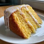 Close-up of a moist, yellow slice of lemon cake recipe topped with white glaze and lemon zest.
