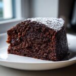 A close-up of a rich, dark slice of gingerbread cake dusted lightly with powdered sugar on a white plate.