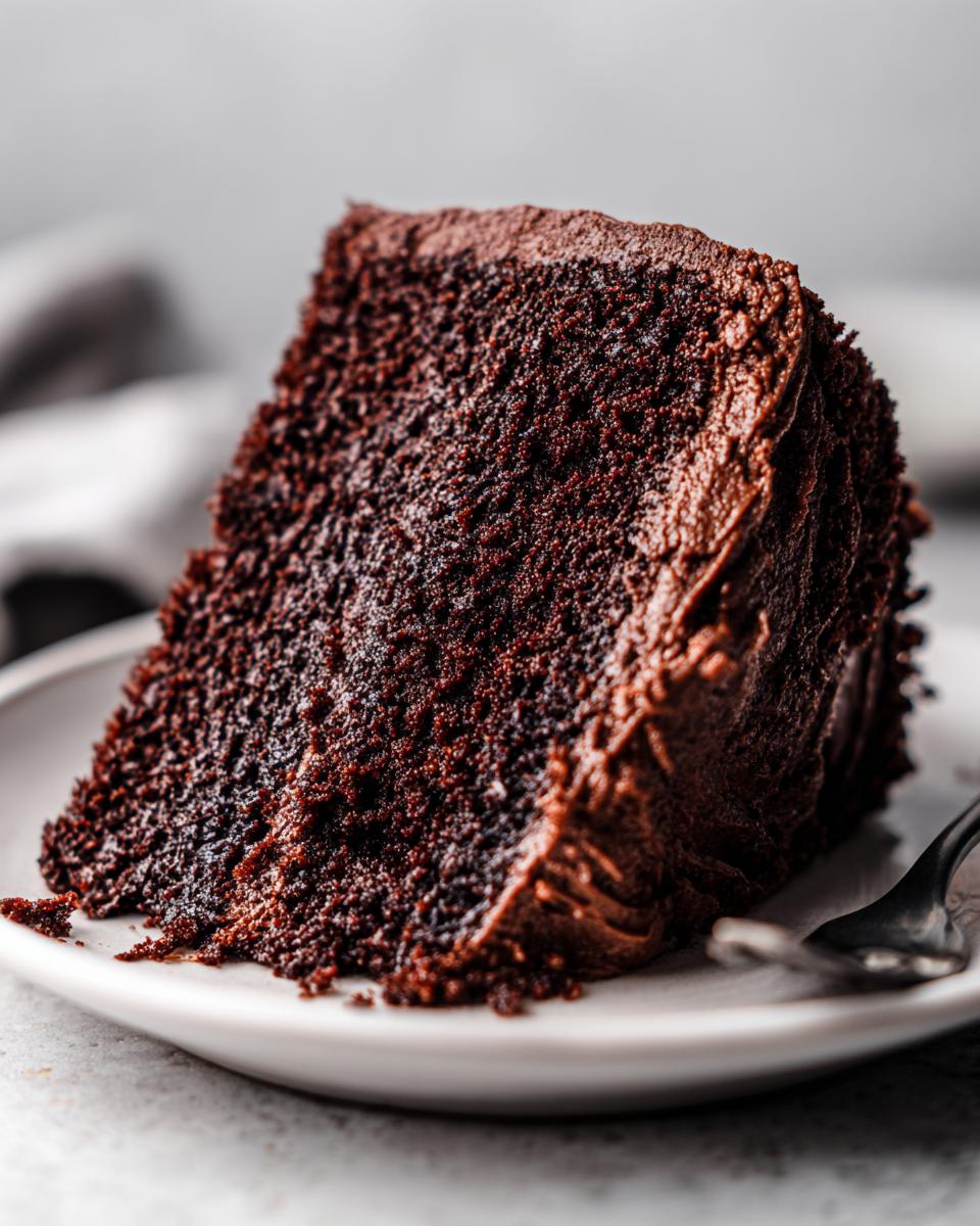Close-up of a rich, dark slice of homemade chocolate cake topped with thick chocolate frosting on a white plate.