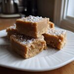 Close-up of several squares of light brown penuche fudge stacked on a white plate, lightly dusted with powdered sugar.