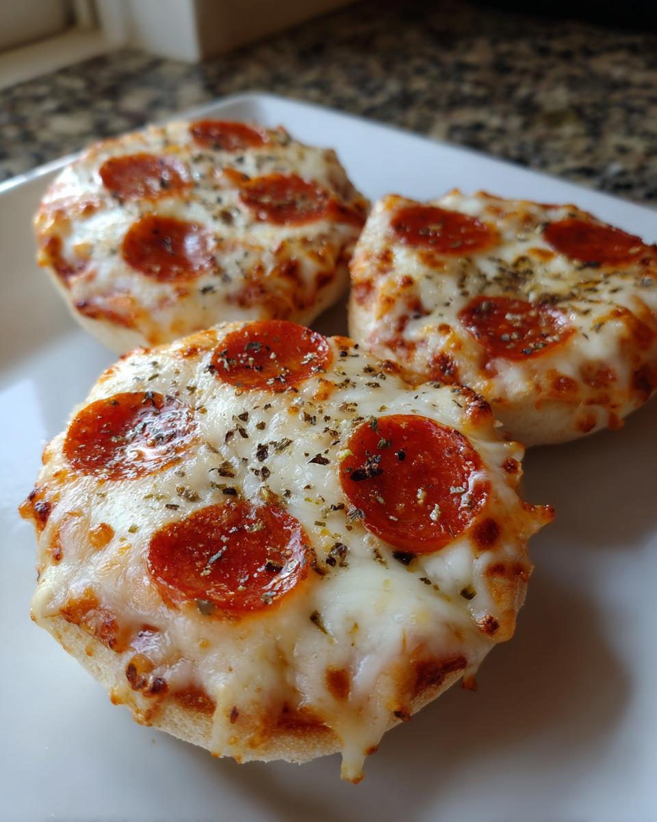 Three freshly baked pepperoni english muffin pizza halves topped with melted cheese and herbs on a white plate.