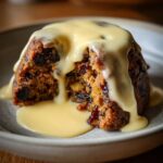 Close-up of a rich, dark plum pudding cut open, generously topped with flowing vanilla custard sauce.
