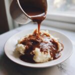A stream of rich brown chicken gravy is being poured from a small pitcher onto a mound of fluffy mashed potatoes.