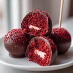Close-up of chocolate-dipped red velvet cake pops, one cut in half showing the bright red interior and cream cheese filling.