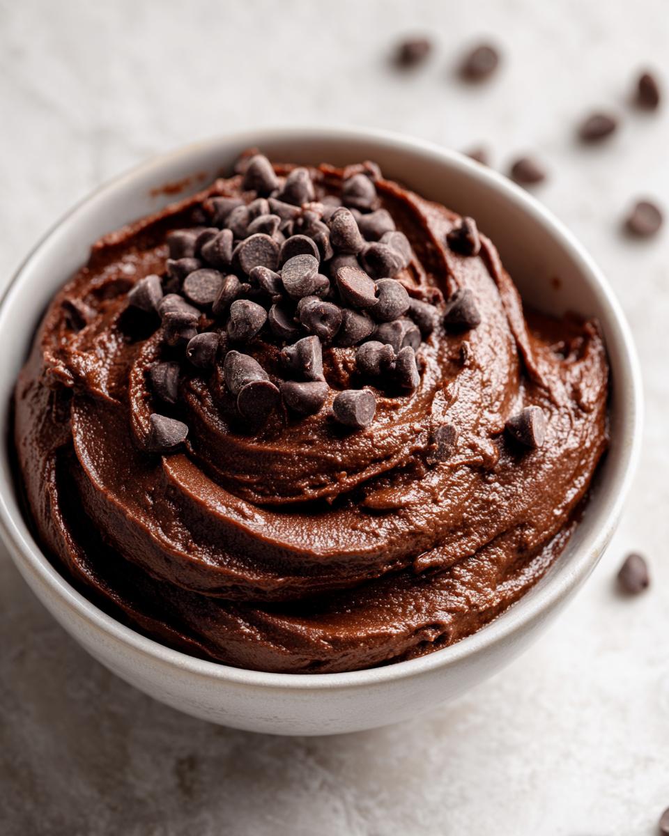 Close-up of rich, dark chocolate brownie batter dip swirled in a white bowl, topped generously with mini chocolate chips.
