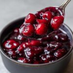 A spoonful of glistening, thick cherry pie filling being lifted from a dark bowl.