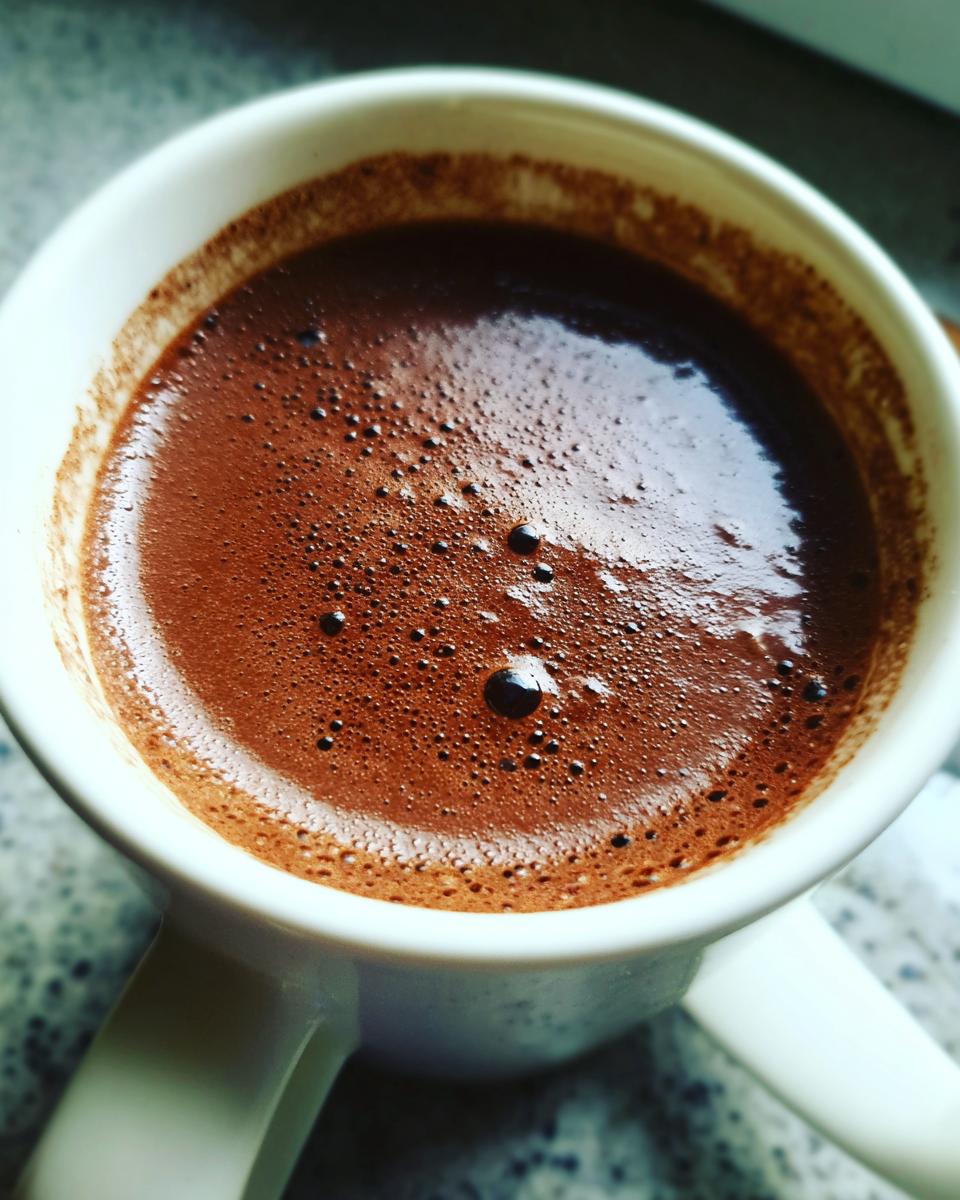 A close-up, overhead view of rich, dark french hot chocolate with a frothy top in a white mug.