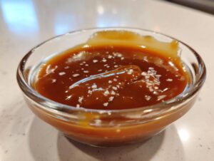 Close-up of rich, brown salted caramel sauce topped with flaky sea salt in a clear glass ramekin.