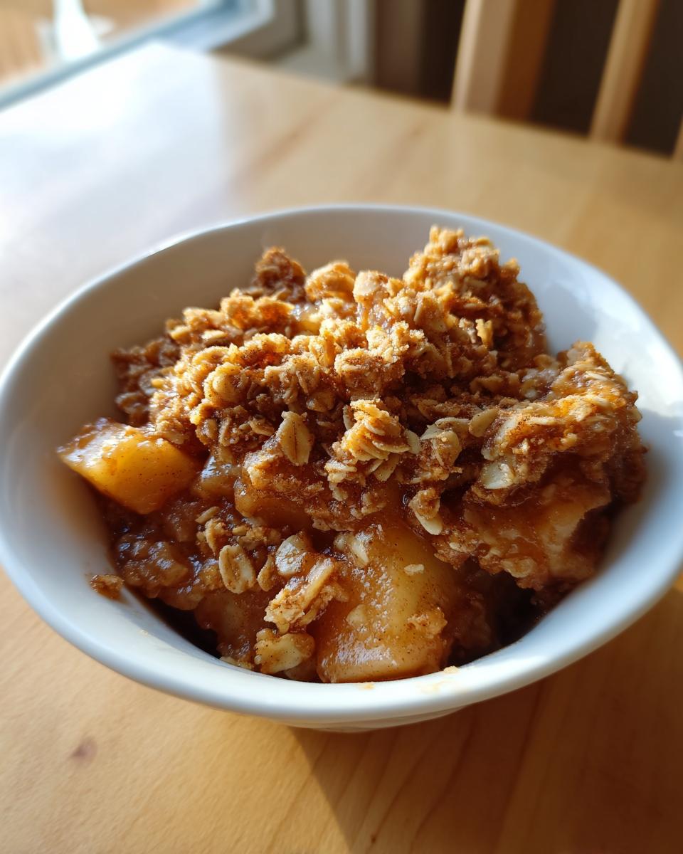 A close-up of a serving of warm apple crisp recipe in a white bowl, featuring baked apples and a crunchy oat topping.
