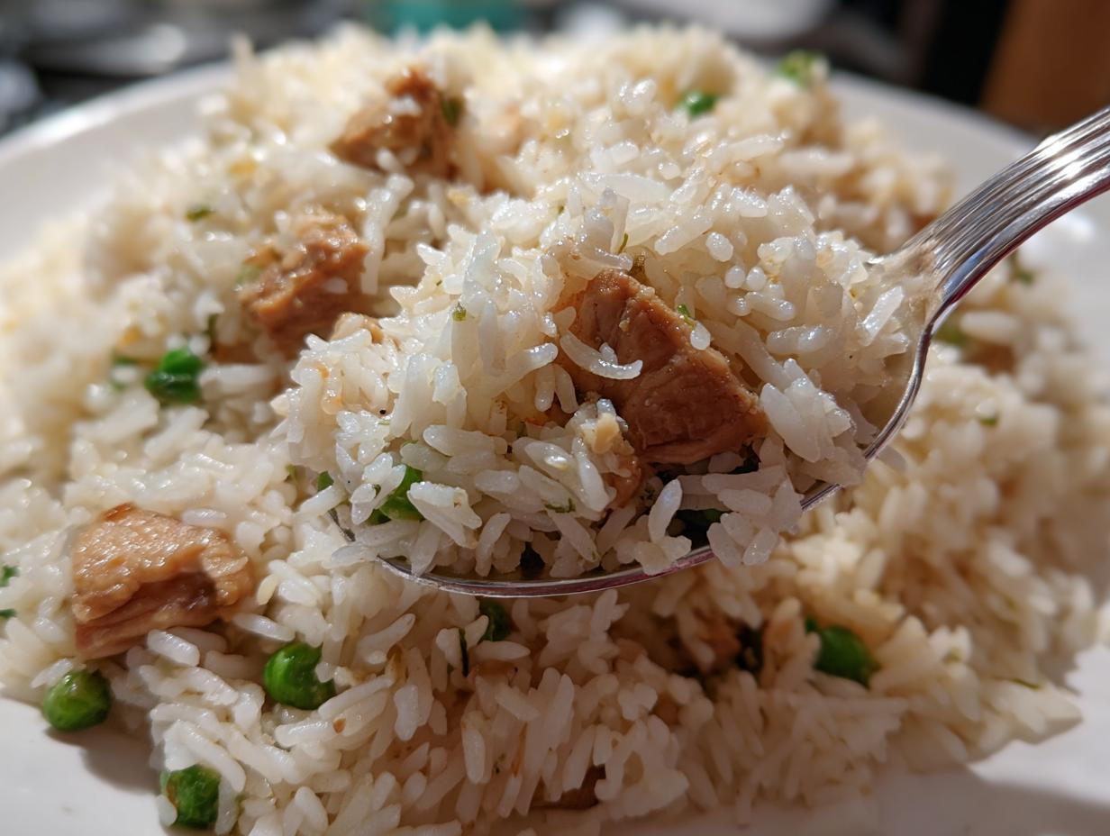 Close-up of a spoonful lifting fluffy white rice mixed with pieces of chicken and green peas from a plate of one pot chicken and rice.