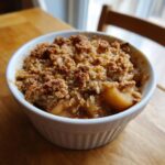 Close-up of a single serving of warm apple crisp recipe in a white ramekin on a wooden table.