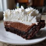 A rich slice of 4 layer mud pie featuring a dark crust, chocolate filling, and a thick layer of whipped cream.
