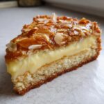 Close-up of a moist slice of bee sting cake showing layers of sponge, rich vanilla cream, and caramelized almond topping.