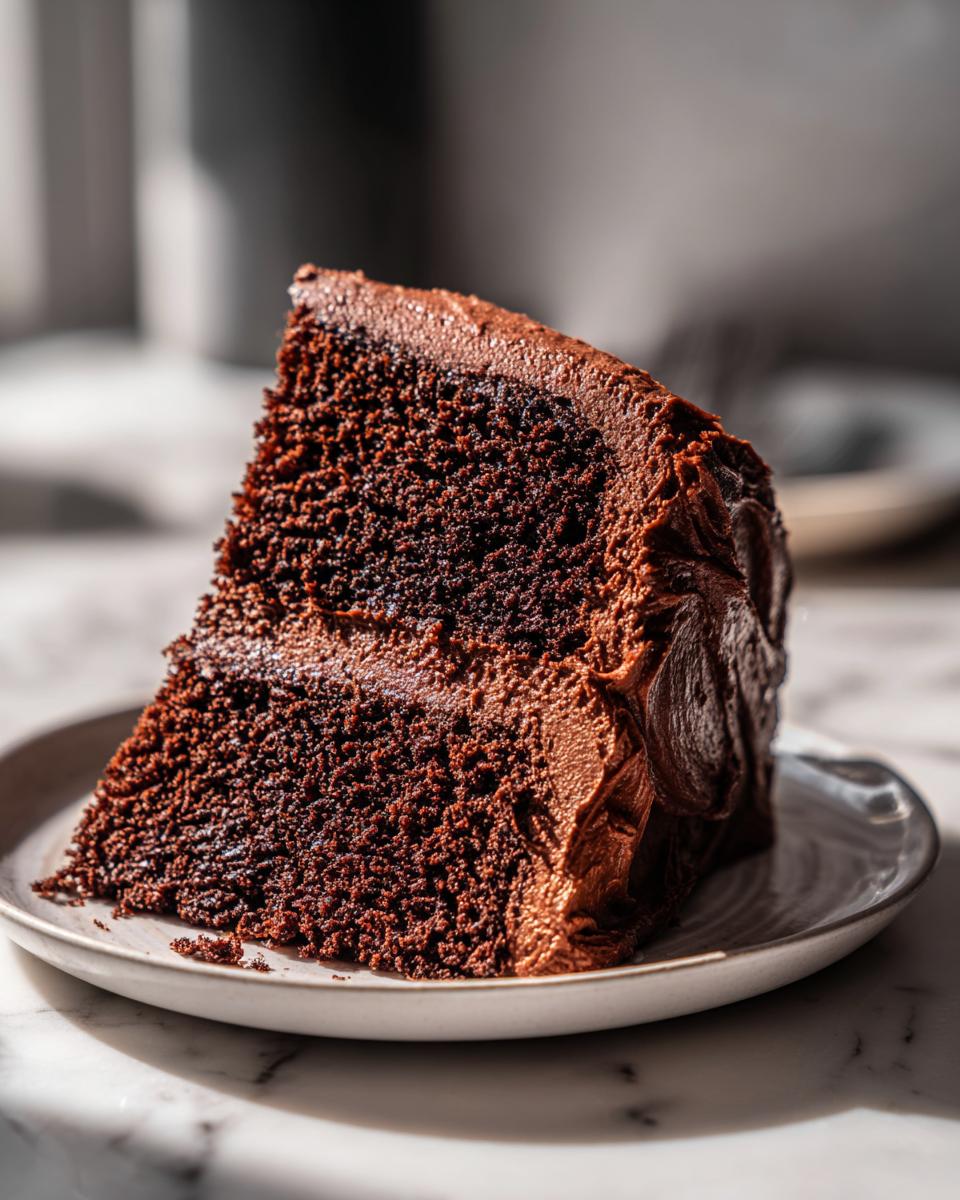 A moist, dark slice of homemade chocolate cake covered in thick chocolate frosting on a small white plate.
