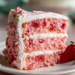 A close-up of a moist slice of three-layer strawberry cake with white and pink frosting, next to a fresh strawberry.