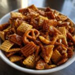 A close-up of a white bowl filled with crunchy, glazed toffee chex mix recipe pieces and pretzels.