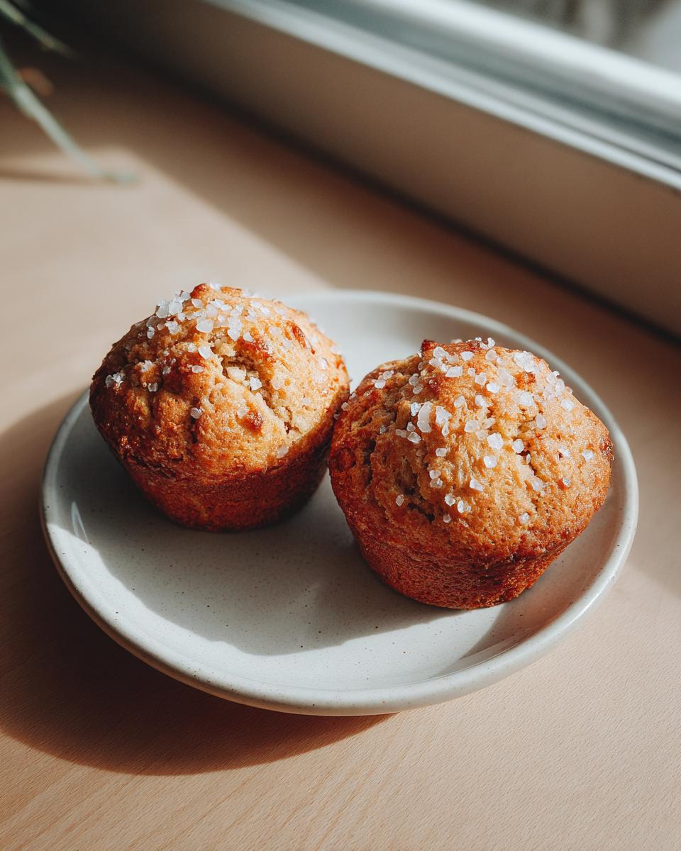 Two golden brown banana oatmeal muffins topped with coarse sugar crystals, sitting on a small speckled plate.
