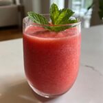 Close-up of a vibrant pink watermelon smoothie in a clear glass, topped with fresh mint leaves.