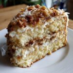 A close-up of a moist slice of white German chocolate cake layered with frosting and topped with coconut and pecans.