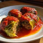 Four delicious cabbage rolls served in a white bowl, covered in rich tomato sauce, with one roll cut open showing the meat filling.