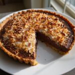 A rich german chocolate pie with a slice removed, topped with toasted coconut and pecans.