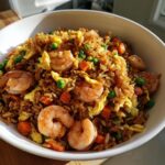 A close-up, appetizing photo of amazing shrimp fried rice mixed with scrambled egg, peas, and diced carrots, served in a white bowl.