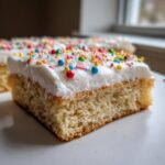 A close-up of a square slice of soft sugar cookie bars topped with thick white frosting and colorful sprinkles.