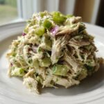 A mound of creamy avocado chicken salad featuring shredded chicken, diced avocado, celery, and red onion on a white plate.