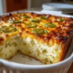A freshly baked popper dip in a white casserole dish with a slice removed, topped with melted cheese and jalapeño slices.