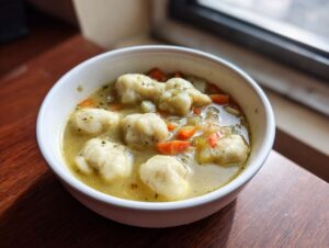 Close-up of a white bowl filled with creamy chicken and dumpling soup, featuring fluffy dumplings and carrots.