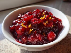 Close-up of bright red homemade cranberry sauce, studded with whole berries and topped with orange zest, served in a white bowl.