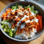 A vibrant buffalo chicken bowl featuring shredded spicy chicken over rice, topped with ranch dressing, lettuce, carrots, and celery.
