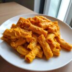 A serving of rigatoni pasta coated thickly in vibrant orange butternut squash pasta sauce, topped with black pepper.