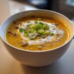 A close-up of a bowl of rich, creamy cheeseburger soup, topped with sour cream and fresh chopped green onions.