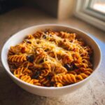 A close-up of a white bowl filled with creamy enchilada pasta, chicken pieces, black beans, and topped with melted shredded cheese.