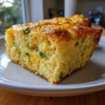 A close-up of a moist slice of mexican cornbread, studded with melted cheese and green peppers, on a white plate.
