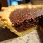A close-up view showing a slice taken out of a rich, dark chocolate chess pie with a flaky crust.