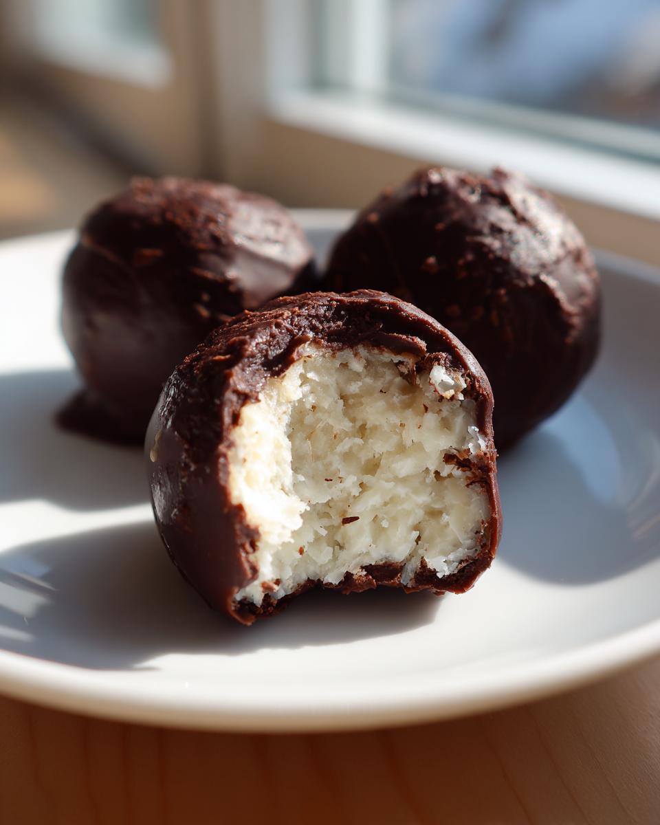 Close-up of chocolate covered coconut balls, one with a bite revealing the shredded white coconut filling.