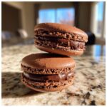 Two perfectly baked chocolate macarons stacked on a granite countertop, showing the ruffled feet and rich chocolate ganache filling.