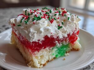 A festive slice of christmas poke cake featuring layers of red and green gelatin, topped with whipped cream and holiday sprinkles.