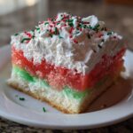 A square slice of colorful Christmas poke cake featuring red and green gelatin layers, whipped cream, and holiday sprinkles.
