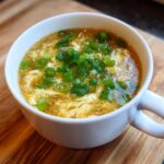 A close-up of a white mug filled with hot egg drop soup, featuring wispy egg ribbons and topped generously with sliced green onions.